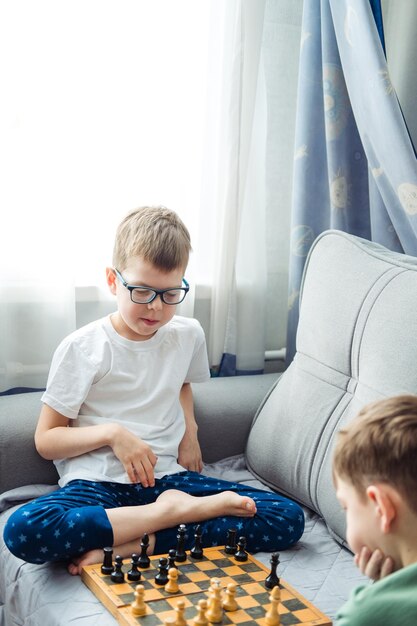 Boys playing wooden chess lying on a gray sofa in front of the window