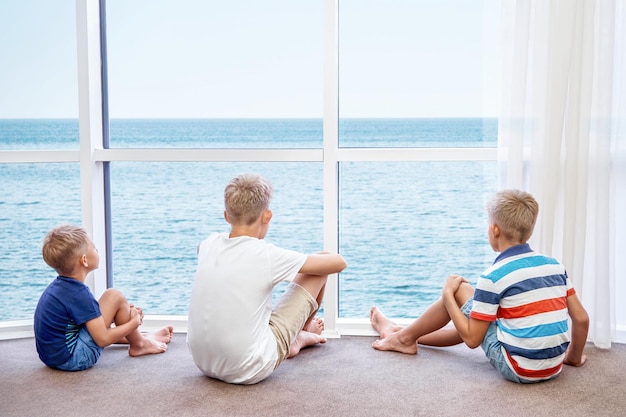 Братья и сестры мальчиков наслаждаются лазурным морем из панорамного окна в гостиничном номере