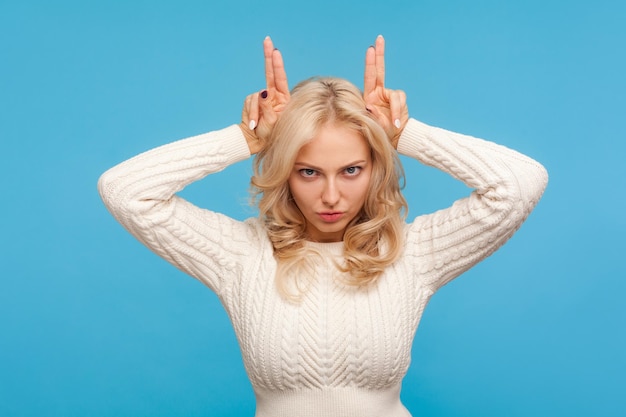 Boze arrogante vrouw met krullend blond haar die vingers boven het hoofd houdt met horens, koppige bazige vrouw die haar humeur toont. Indoor studio-opname geïsoleerd op blauwe achtergrond