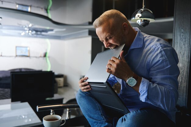 Boze blanke man van middelbare leeftijd, ondernemer in vrijetijdskleding zittend op een tafel met een kopje koffie en een laptop in zijn handen, erop bijtend van woede