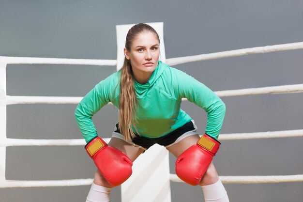 Boze boksen atletische jonge vrouw in groene lange mouw in evenwicht met rode bokshandschoenen en kijken naar camera met serieus gezicht. indoor studio opname op grijze achtergrond.