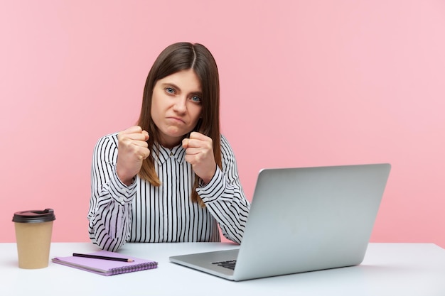 Boze geërgerde vrouw in gestreept shirt zittend op laptop op de werkplek met gebalde vuisten om te slaan, met boksgebaar, klaar om te vechten voor een baan. Indoor studio-opname geïsoleerd op roze background