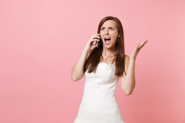 Boze, geïrriteerde vrouw in witte jurk spreidt handen, praat op mobiele telefoon, schreeuwt en vloekt