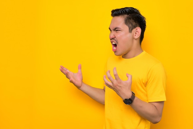 Boze jonge Aziatische man in casual tshirt hand opsteken en schreeuwen met kopie ruimte geïsoleerd op gele achtergrond Mensen lifestyle concept