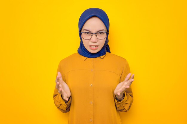 Boze jonge Aziatische moslimvrouw gekleed in een oranje shirt die handen opsteekt en luid schreeuwt, drukt woede uit geïsoleerd op gele achtergrond