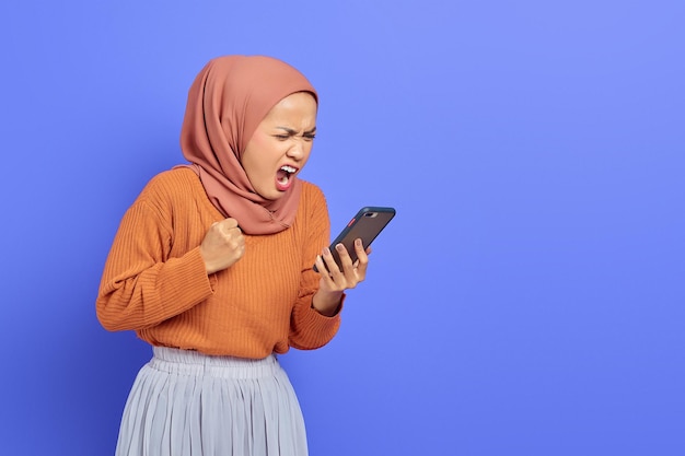 Boze jonge Aziatische vrouw in bruine trui en hijab die smartphone gebruikt terwijl ze luid schreeuwt geïsoleerd over paarse achtergrond. Mensen islam religieus concept