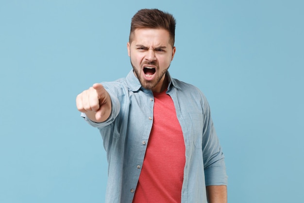 Boze jonge bebaarde man in casual shirt poseren geïsoleerd op pastel blauwe achtergrond studio portret. Mensen levensstijl concept. Bespotten kopie ruimte. Wijsvinger op camera wijzen, vloekend schreeuwend.