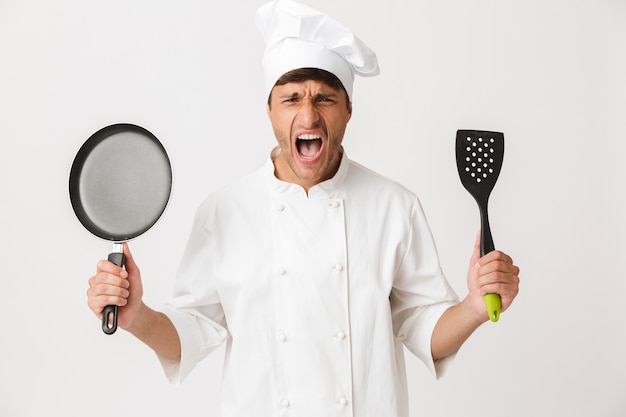 Boze jonge chef-kok man staande geïsoleerd op een witte muur met koekenpan