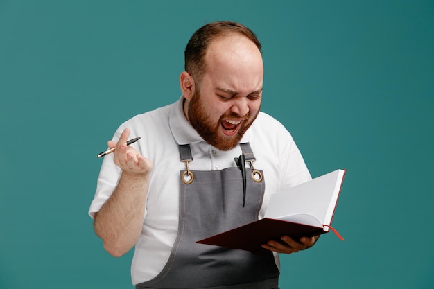 Foto boze jonge mannelijke kapper met wit overhemd en kappersschort met pen en notitieblok kijken naar notitieblok schreeuwen geïsoleerd op blauwe achtergrond