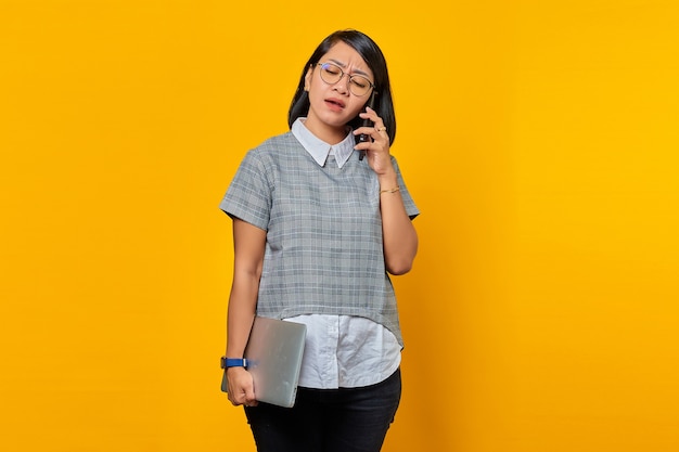 Boze jonge vrouw die op smartphone spreekt en laptop op gele achtergrond houdt