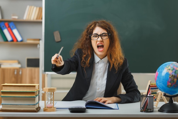 boze jonge vrouwelijke leraar die een bril draagt, wijst naar de camera met de aanwijzer aan het bureau met schoolhulpmiddelen in de klas