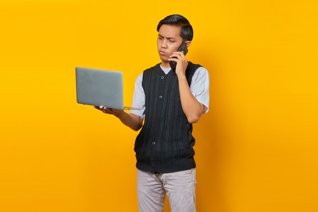 Boze knappe jonge man praten op smartphone en kijken naar laptop scherm op gele achtergrond