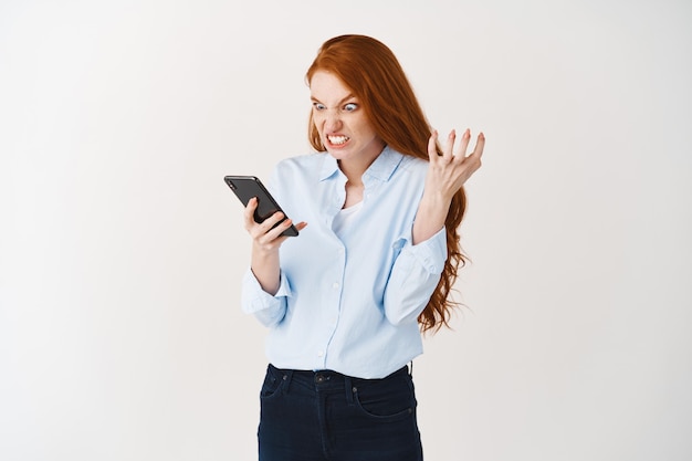 Boze roodharige vrouw die pissig kijkt naar het smartphonescherm en woedend grimast, knijpt in de hand van woede en staart naar de telefoon, witte muur