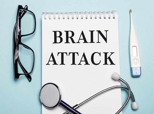 Brain attack scritto su un blocco note bianco accanto a uno stetoscopio, occhiali e un termometro elettronico su sfondo azzurro