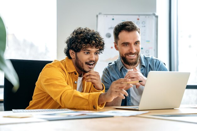 Photo brainstorming excited satisfied male colleagues working together in a creative office using laptop discussing a new project planning deadlines prospects and promotion strategy smile