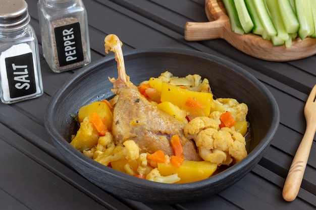 Braised domestic duck with vegetables in a black clay plate standing on wooden background