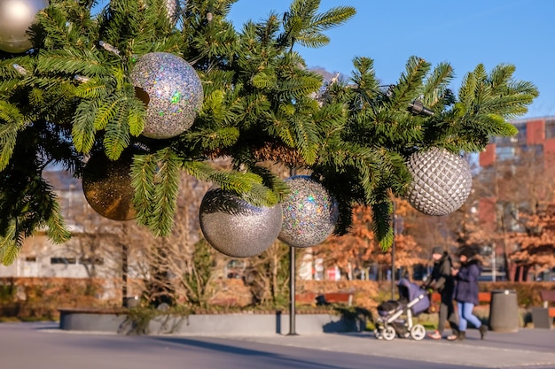 Branch of artificial Christmas tree with white christmas balls on city street outdoors at sunny winter day. Blurred family with child walking.