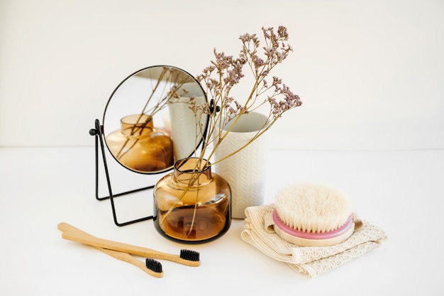 Foto ramo in un vaso di vetro, specchio, spazzolino da denti in bambù e prodotti cosmetici sul tavolo bianco. arredamento per interni. decorazione alla moda per la casa.