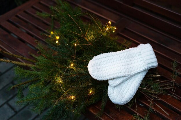 Branches of a coniferous tree decorated with a garland lie on a bench along with knitted white mittens