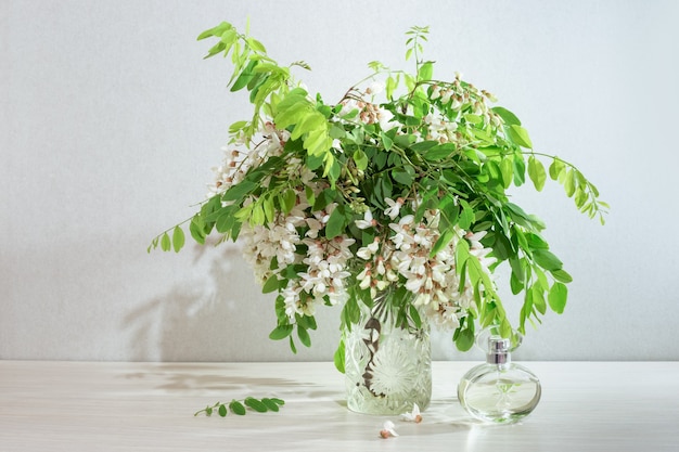 Branches with white acacia flowers in a transparent glass vase, next to a bottle of perfume. Spring scents of nature.