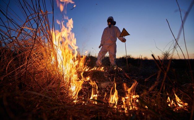 Brandweerman ecoloog die's nachts vuur bestrijdt in het veld Man in beschermend stralingspak en gasmasker bij brandend gras met rook Natuurlijke rampconcept