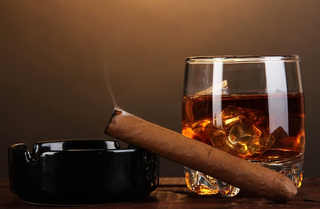 Brandy glass with ice and cigar on wooden table on brown surface