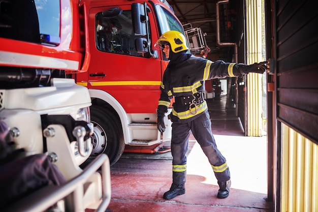 Coraggioso giovane vigile del fuoco in uniforme protettiva con casco sulla testa in piedi nella stazione dei vigili del fuoco e apertura della porta