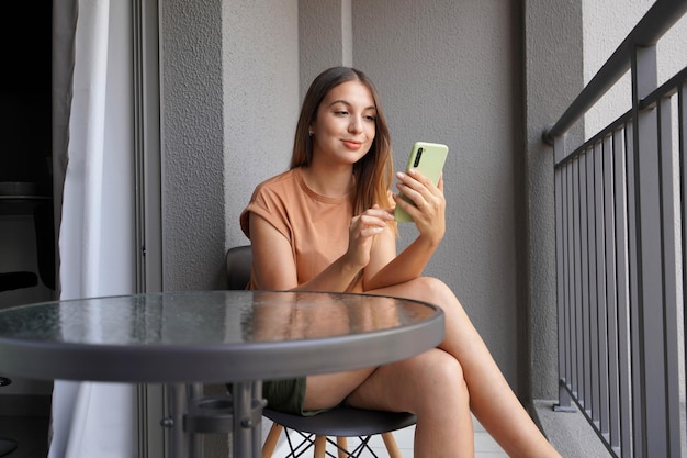 Foto braziliaans meisje gebruikt smartphone om te chatten of berichten te controleren ontspannen op haar balkon thuis
