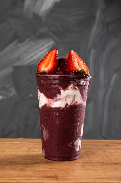 Braziliaanse Frozen AÃ§ai Berry Ice Cream Smoothie in plastic beker met strobessen en gecondenseerde melk. Op een houten bureau en een grijze zomerachtergrond. Vooraanzicht voor menu en sociale media