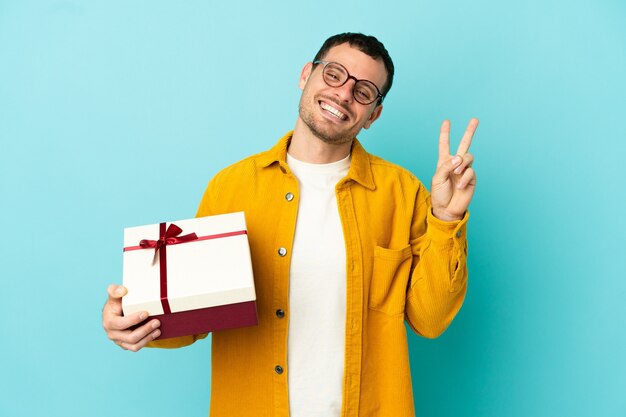 Braziliaanse man met een geschenk over een geïsoleerde blauwe achtergrond die lacht en een overwinningsteken toont