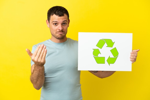 Braziliaanse man over geïsoleerde paarse achtergrond met een bordje met recycle-pictogram en komend gebaar doen