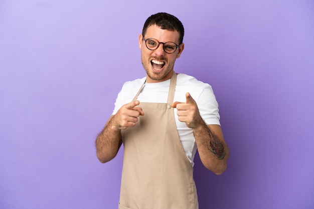 Braziliaanse restaurantkelner over geïsoleerde paarse achtergrond die naar voren wijst en glimlacht