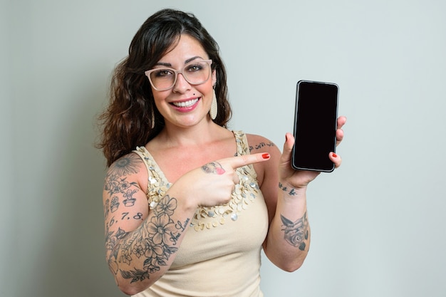 Braziliaanse vrouw met een bril, getatoeëerd, glimlachend en wijzend met haar vinger naar smatphone.