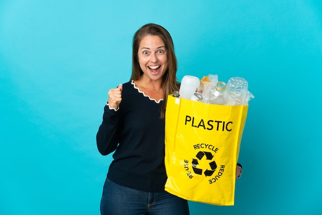 Braziliaanse vrouw van middelbare leeftijd die een zak vol plastic flessen houdt om op blauwe muur te recyclen die een overwinning in winnaarspositie viert