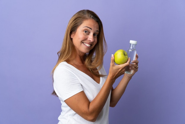 Braziliaanse vrouw van middelbare leeftijd geïsoleerd op paarse muur met een appel en met een fles water