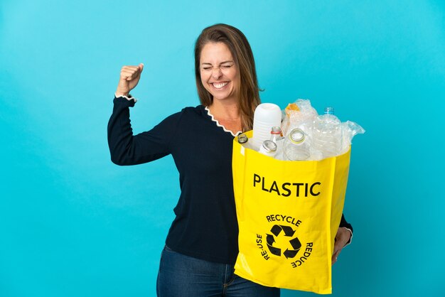 Braziliaanse vrouw van middelbare leeftijd met een zak vol met plastic flessen om te recyclen op blauw doet sterk gebaar