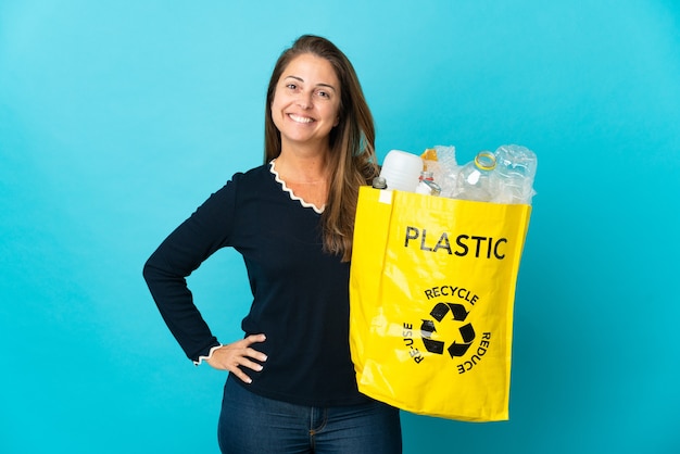 Braziliaanse vrouw van middelbare leeftijd met een zak vol plastic flessen om te recyclen op blauwe muur poseren met armen op heup en glimlachen