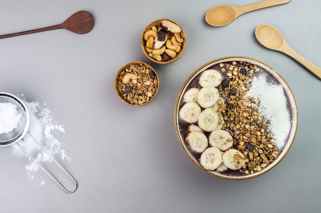 Brazilian acai in a white bowl with banana powdered milk granola and nuts in small bamboo bowls Cashew nuts and Para Top view Grey background Selective focus