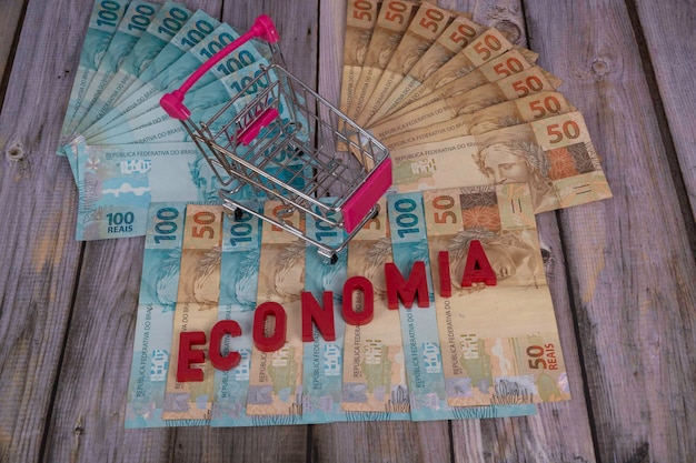 Brazilian banknotes Banknotes of 50 and 100 reais in the background with the word economia in Portuguese economy in English in red with a mini supermarket cart Selective focus