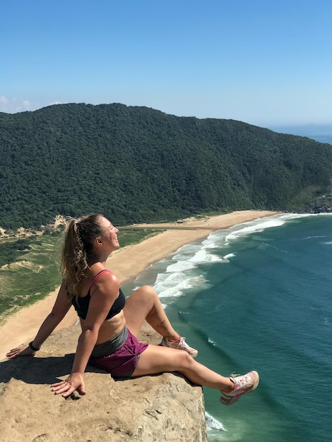 Photo brazilian blonde woman sitting on a rock on the cliff in florianopolis santa cartarina lagoinha do leste beach background