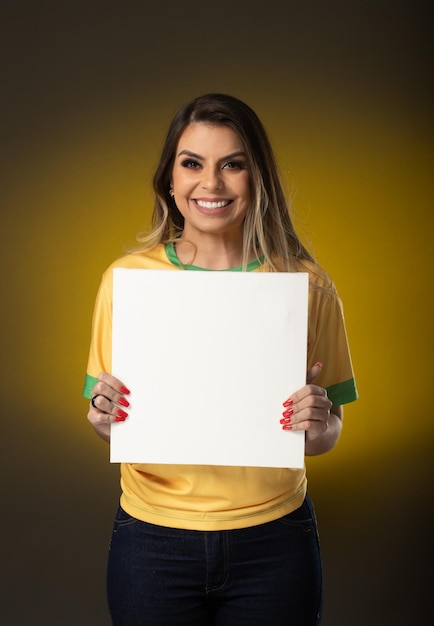 Ventola brasiliana holding white blank card donna fan che celebra in una partita di calcio o di calcio su sfondo giallo colori del brasile