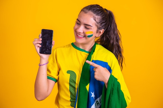 Ritratto del fan brasiliano fan brasiliano che mostra il suo telefono cellulare vestito come un tifoso di calcio o una partita di calcio su sfondo giallo colori della coppa del mondo del brasile