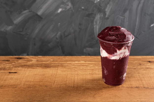 Brazilian Frozen AÃ§ai Berry Ice Cream Smoothie in plastic cup with Condensed Milk. On a wooden desk and a gray summer background. Front view for menu and social media