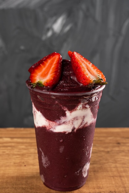 Brazilian Frozen AÃ§ai Berry Ice Cream Smoothie in plastic cup with Straw Berries and Condensed Milk. On a wooden desk and a gray summer background. Front view for menu and social media