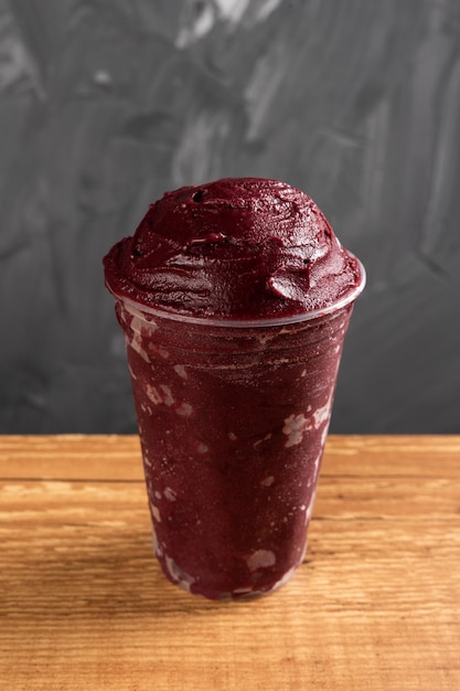 Brazilian Frozen AÃ§ai Berry Ice Cream Smoothie in plastic cup. On a wooden desk and a gray summer background. Front view for menu and social media