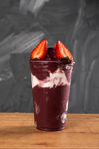 Brazilian Frozen AÃ§ai Berry Ice Cream Smoothie in plastic cup with Straw Berries and Condensed Milk. On a wooden desk and a gray summer background. Front view for menu and social media