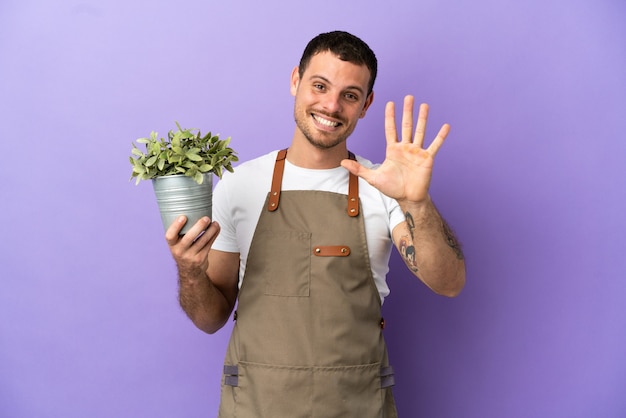 指で5を数える孤立した紫色の背景の上に植物を保持しているブラジルの庭師の男