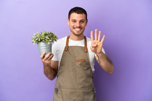 幸せな孤立した紫色の背景の上に植物を保持し、指で4を数えるブラジルの庭師の男