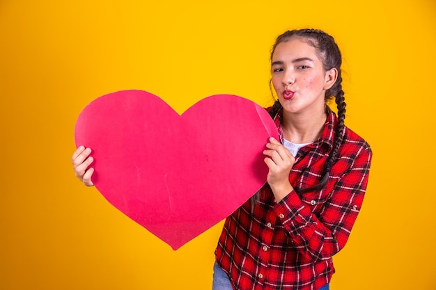 Photo brazilian girl wearing typical clothes for the festa junina holding in her hands a paper banner in the shape of a heart concept of tent of the kiss june party and june party