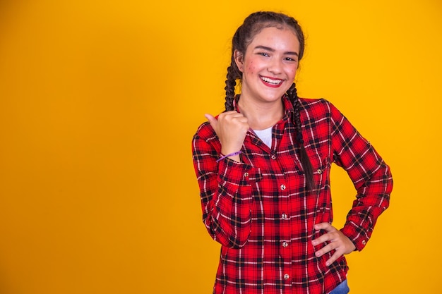 Brazilian girl wearing typical clothes for Festa Junina pointing with thumb to the free side pro text